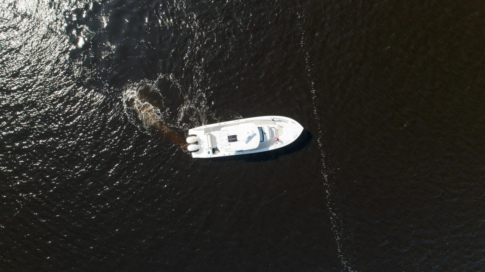 A white boat and its swirling wake seen directly from above on sunlit water.