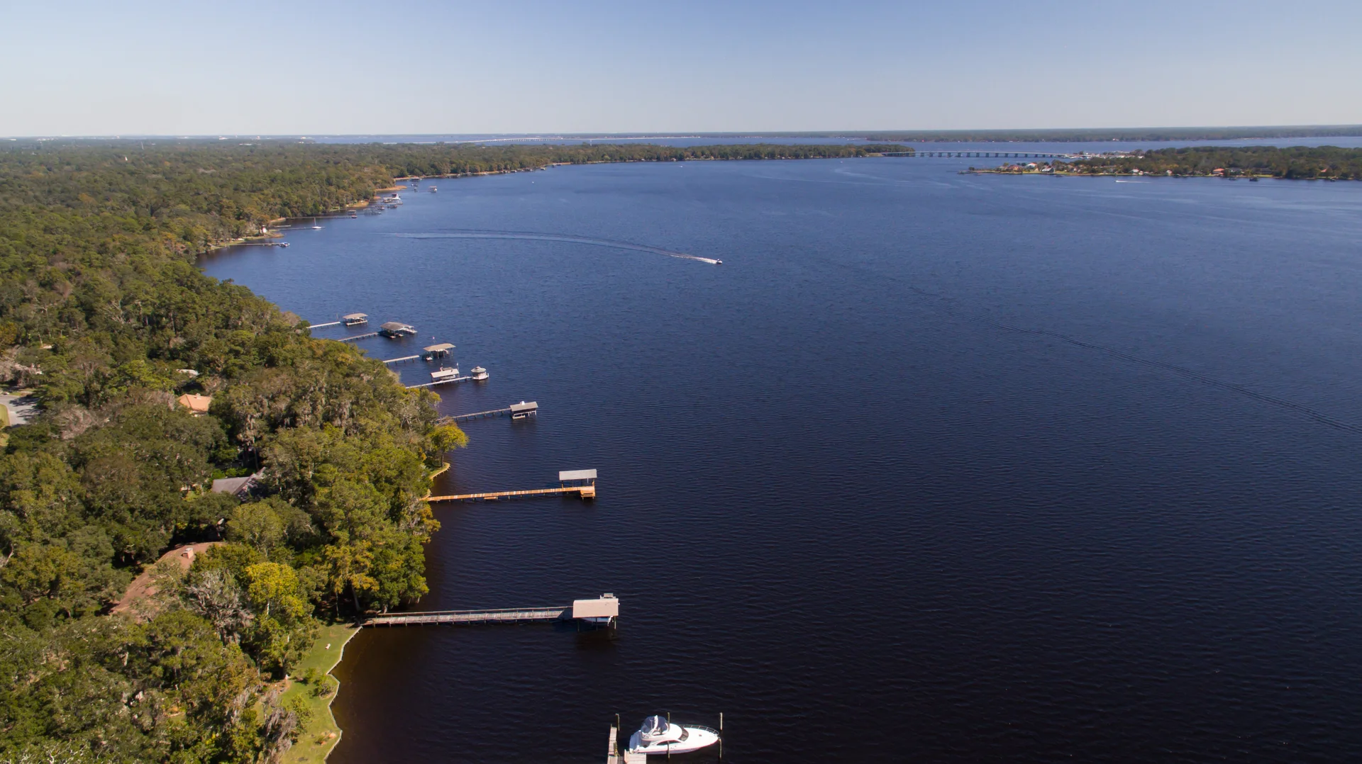 An aerial view of a wooded shoreline, docks and a boat crossing the river.